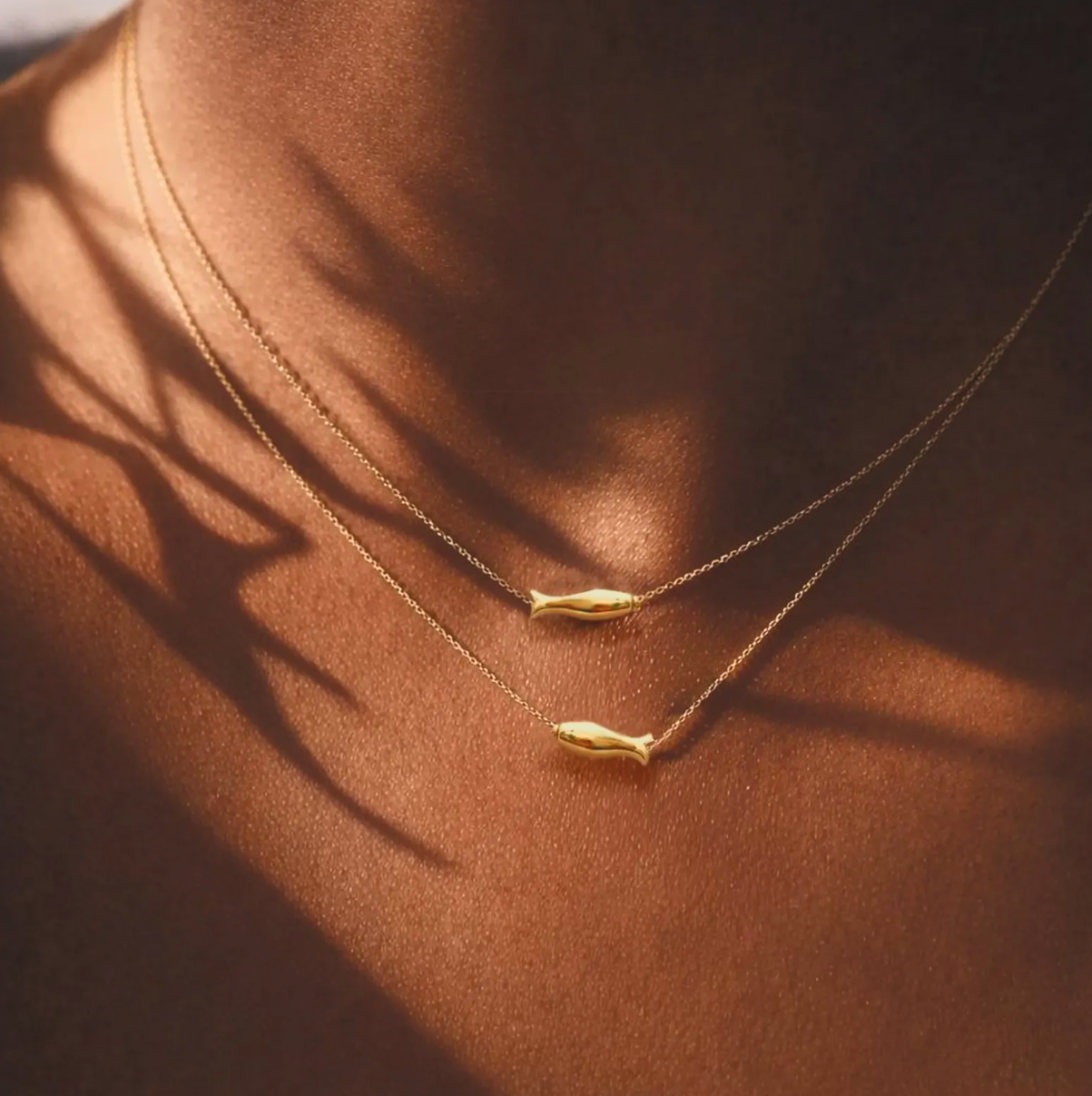 Dainty layered gold fish necklace on sun-kissed skin with artistic shadow, coastal summer jewelry by LP-Muse.